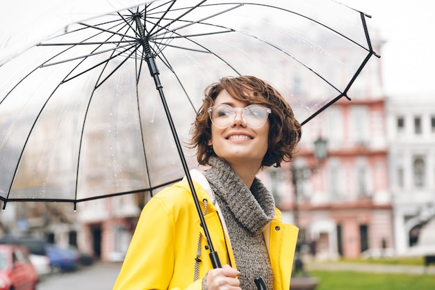 Verbazend portret van gelukkige vrouw in gele regenjas die in stad onder transparante paraplu tijdens koude regenachtige dag lopen