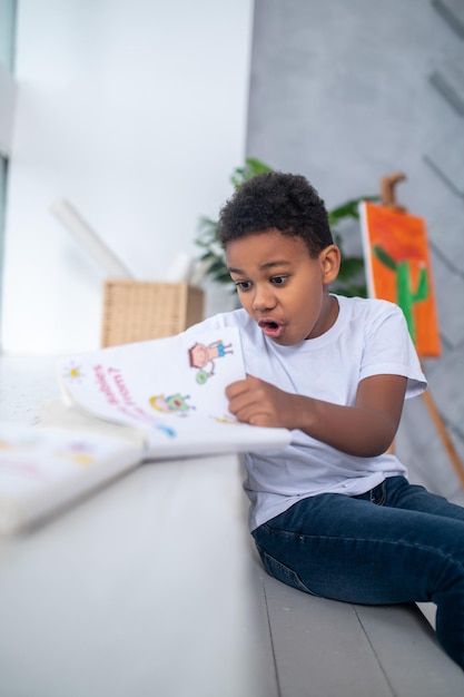 Verbazing. Donkere schoolgaande jongen in wit t-shirt kijkt verbaasd naar boek met open mond zittend in een lichte kamer bij daglicht