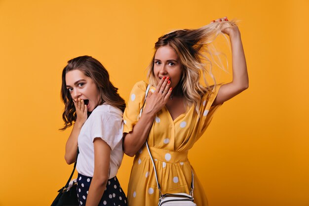 Verbazingwekkende bruinharige meisje verrast emoties uiten terwijl poseren met beste vriend. Blanke blonde vrouw met plezier met zus tijdens portretfotografie.