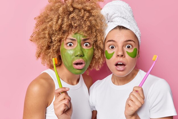 Verbijsterd twee vrouwen houden tandenborstels zorgen voor mondhygiëne en schoonheidsprocedures passen een groen gezichtsmasker en schoonheidspleisters toe, gekleed in casual witte t-shirts geïsoleerd over roze studiomuur