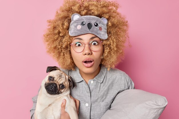 Verbijsterde krullende vrouw staart afgeluisterde ogen en geopende mond kan niet geloven in schokkend nieuws houdt pug dog en zacht kussen gekleed in nachtkleding poses binnen Mensen dieren en slaaptijd concept