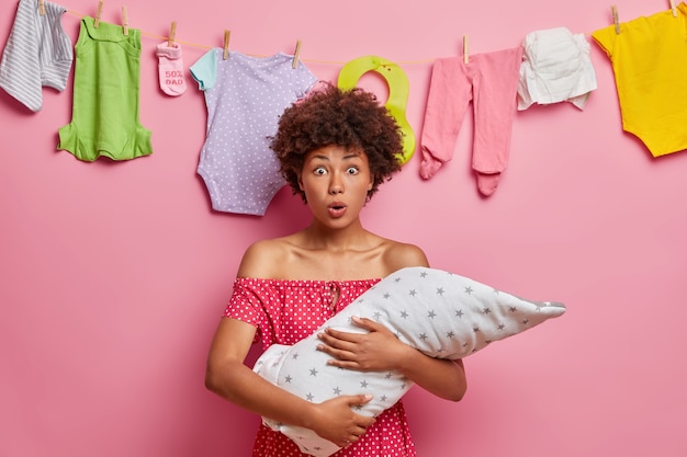 Verbijsterde moeder met krullend haar houdt baby vast, komt te weten over de ernstige ziekte van het kind, houdt de mond wijd open, poseert