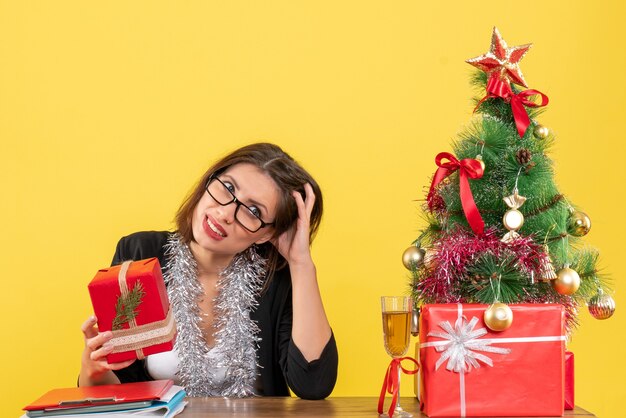 Verbijsterde zakelijke dame in pak met bril met haar cadeau en zittend aan een tafel met een kerstboom erop in het kantoor