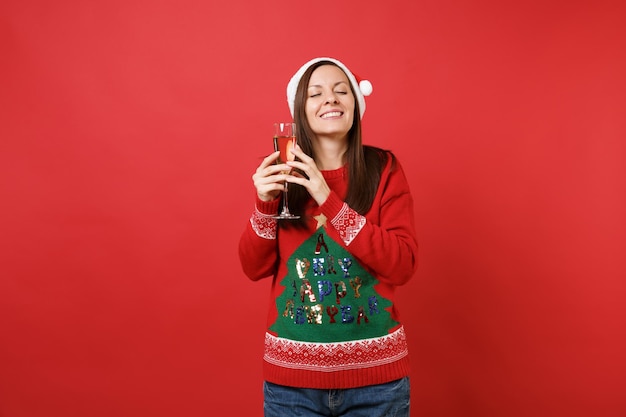 Verbluffend jong santa-meisje in een kerstmuts die de ogen gesloten houdt, met een glas champagne geïsoleerd op een heldere rode achtergrond. gelukkig nieuwjaar 2019 viering vakantie partij concept. bespotten kopie ruimte.