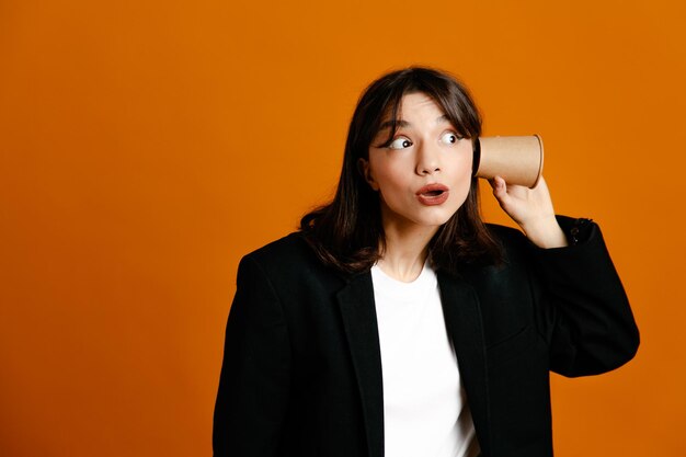 Verdachte bedrijf koffiekopje op oor jonge mooie vrouw draagt zwarte jas geïsoleerd op oranje achtergrond