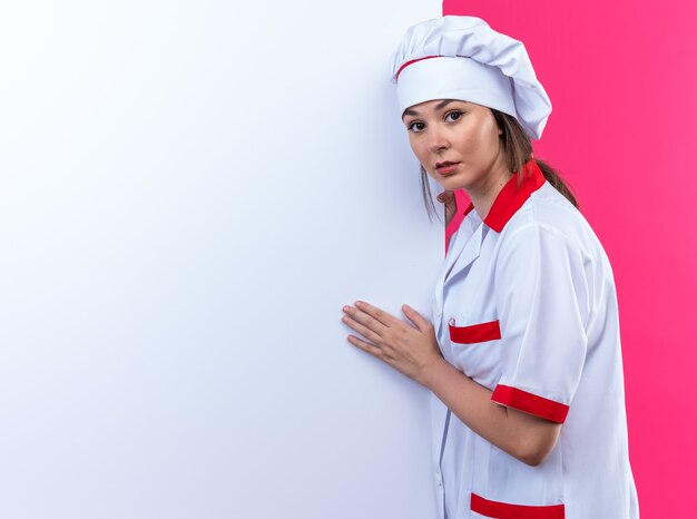 Verdachte jonge vrouwelijke kok in uniform van de chef-kok staat in de buurt van een witte muur geïsoleerd op een roze achtergrond met kopieerruimte