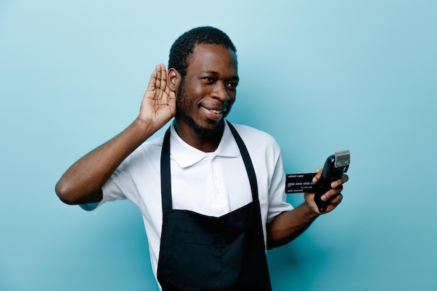 Verdachte tonen luisteren gebaar houden kaart met tondeuses jonge Afro-Amerikaanse kapper in uniform geïsoleerd op blauwe achtergrond