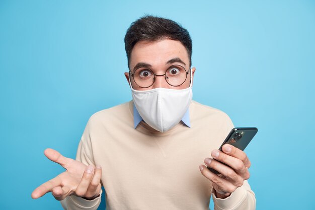 Verdoofde beschaamde volwassen man staart geschokt, draagt masker om besmetting door coronavirus te voorkomen blijft veilig tijdens thuisquarantaine gebruikt mobiele telefoon