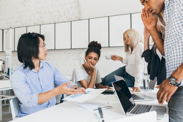 Verdrietig Afrikaans meisje vond fout in documenten en dacht aan iets terwijl haar collega's probeerden te helpen. Aziatische web-ontwikkelaar houdt rapport en legt het uit aan Europese jonge vrouw.