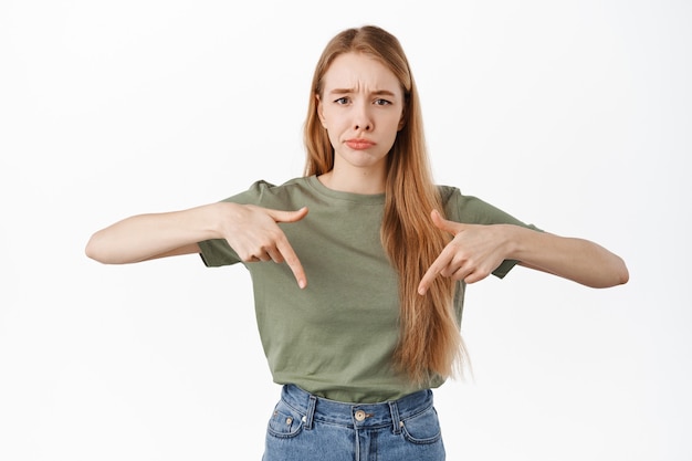 Gratis foto verdrietig en somber meisje dat met de vingers naar beneden wijst, mokkend en fronsend naar oneerlijke verontrustende situatie, klagen, jaloers of teleurgesteld voelen, over witte muur staan