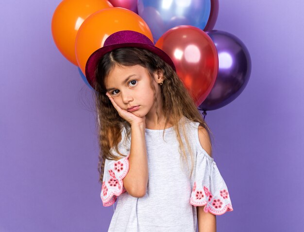 verdrietig Kaukasisch meisje met violet feestmuts hand zetten gezicht staande voor helium ballonnen geïsoleerd op paarse muur met kopie ruimte