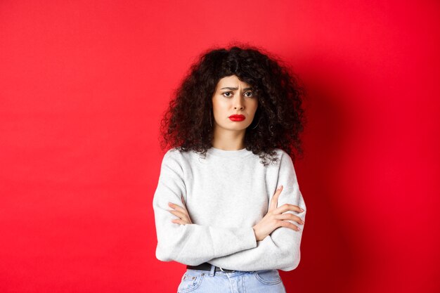 Verdrietige en bezorgde blanke vrouw fronst, kruist de armen op de borst en kijkt bezorgd, voelt zich slecht, staat op een rode achtergrond