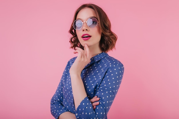 Verfijnd kaukasisch meisje in paarse zonnebril poseren met geïnteresseerde gezichtsuitdrukking jocund jonge vrouw met donker haar chillen tijdens fotoshoot op roze achtergrond