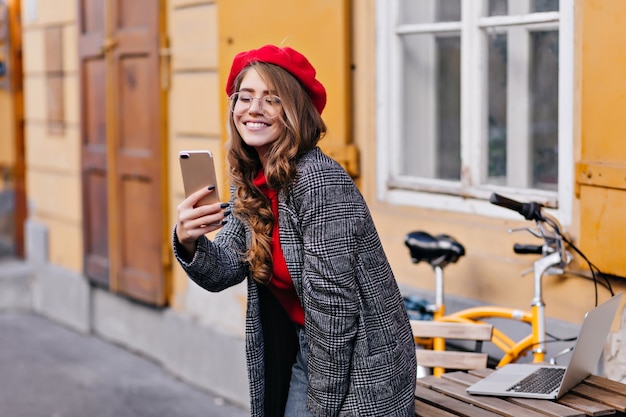Verfijnd krullend Frans meisje selfie maken in de buurt van houten tafel met laptop erop
