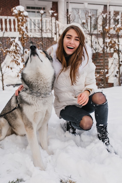 Verfijnd vrouwelijk model in warme kleren die tijdens de wintervakantie met een husky hond dollen. Buitenportret van prachtige jonge dame speelt met huisdier in de ochtend van december.