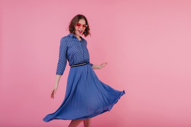 Verfijnd wit meisje met golvend haar dat op roze muur danst. innemende europese dame draagt blauwe midi-rok en blouse.