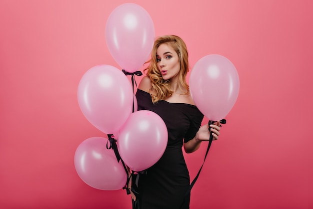 Verfijnde blanke vrouw met ballonnen poseren met verbaasde gezichtsuitdrukking. portret van aantrekkelijk krullend meisje dat op verjaardagsfeestje wacht.