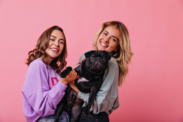 Verfijnde bruinharige meisje poseren met plezier naast schattige puppy. Elegante blonde jonge vrouw met zwarte bulldog op rooskleurig.