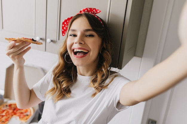 Verfijnde dame met golvend haar selfie maken tijdens het diner. Binnen schot van zalig Kaukasisch meisje dat pizza eet.