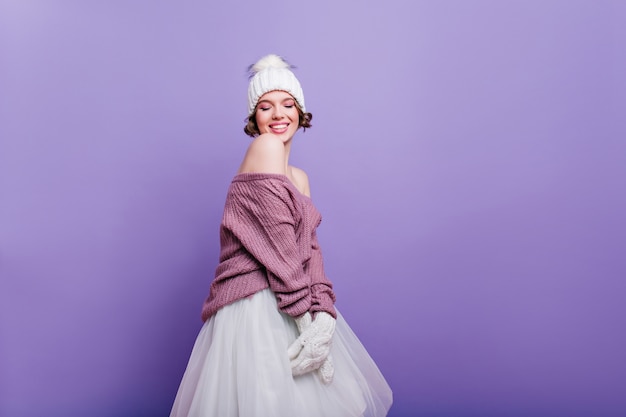 Verfijnde europese vrouw in witte hoed en rok poseren met geïnspireerde gezichtsuitdrukking. trendy korthaar meisje in trui geïsoleerd op paarse muur.