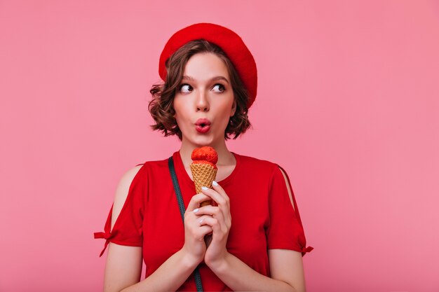 Verfijnde jonge vrouw die in glamoureuze baret ijs eet. emotioneel meisje in rode outfit genieten van dessert.