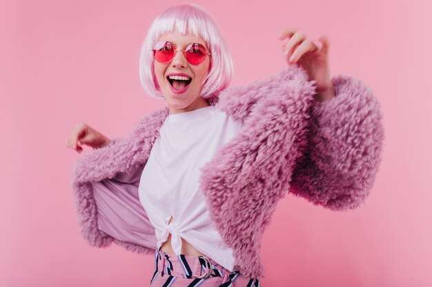 Verfijnde jongedame in korte periwig dansen op roze muur. Schattig meisje lachen in zonnebril poseren in stijlvolle bontjas