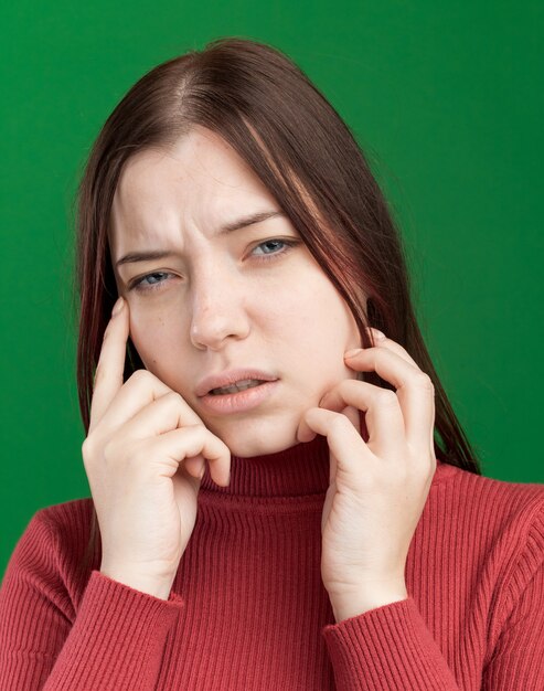 Vergrote weergave van een verwarde jonge mooie vrouw die naar de voorkant kijkt en haar gezicht aanraakt en een denkgebaar doet geïsoleerd op een groene muur