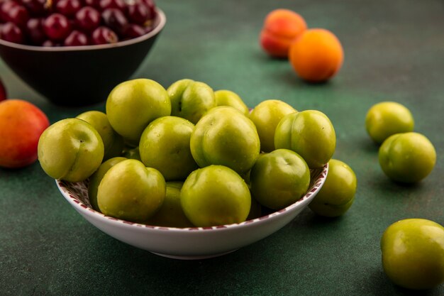 Vergrote weergave van groene pruimen in kom met kom kersen en abrikozen op groene achtergrond