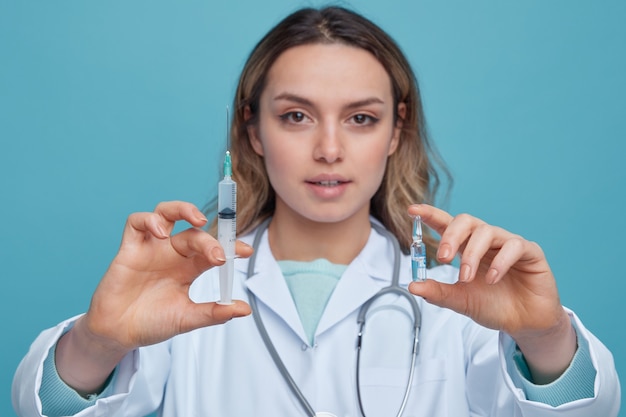 Vergrote weergave van zelfverzekerde jonge vrouwelijke arts die medische mantel en stethoscoop om nek draagt die spuit en ampul naar camera uitstrekt