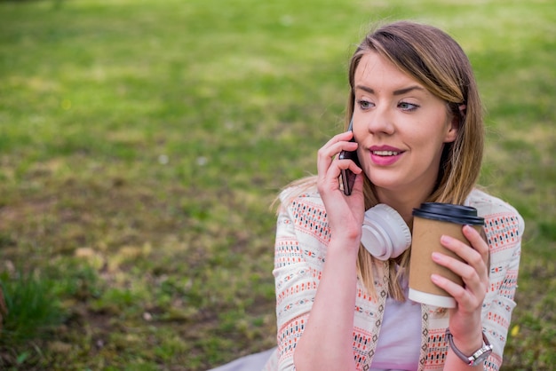 Verheugde vrouw die in de buitenlucht in gesprek zit. Meisje met koffie drinken praten op smartphone. Vrouw in koptelefoon luisteren muziek in de natuur