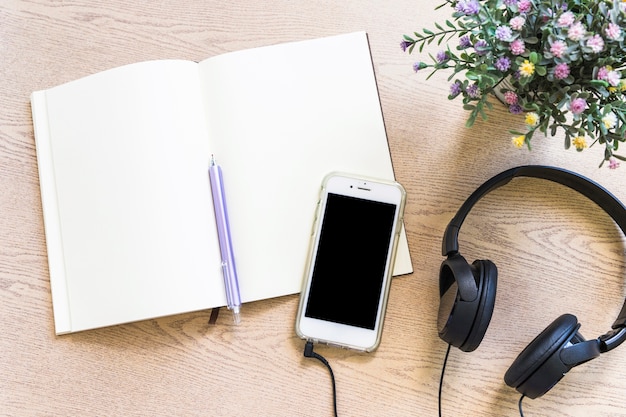 Verhoogde mening van leeg boek met pen; mobiele telefoon en oortelefoon op houten tafel