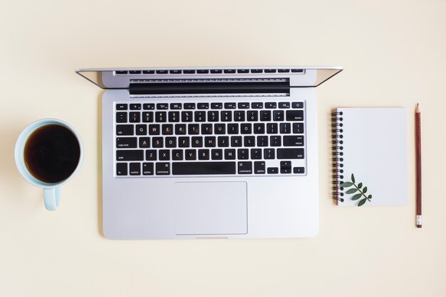 Verhoogde weergave van geopende laptop; kopje thee; spiraal blocnote en potlood op beige achtergrond