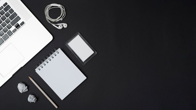 Verhoogde weergave van laptop; oortelefoons; verfrommeld papier; potlood; lege spiraal blocnote en frame op zwarte achtergrond