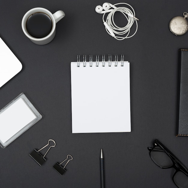 Verhoogde weergave van oortelefoon; koffiekop; paperclips; lenzenvloeistof; met lege witte kladblok gerangschikt op zwarte achtergrond