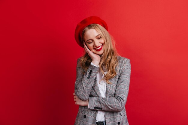 Verlegen blond meisje in baret glimlachend op rode achtergrond Studio shot van modieuze blanke vrouw in jasje