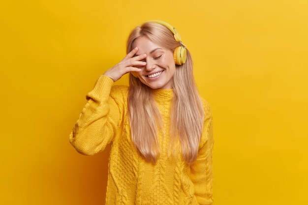 Verlegen positieve blonde vrouw glimlacht in grote lijnen sluit ogen geniet van het luisteren naar favoriete muziek via draadloze koptelefoon besteedt vrije tijd alleen met aangename liedjes draagt trui