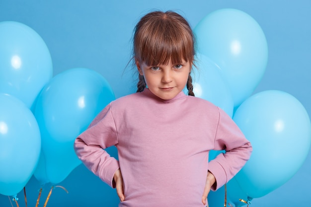 Verlegen schattig klein kind meisje poseren met blauwe lucht ballonnen geïsoleerd over kleur achtergrond. Mooie jongen camera van onder voorhoofd kijken, handen op de heupen houden, roze trui dragen.