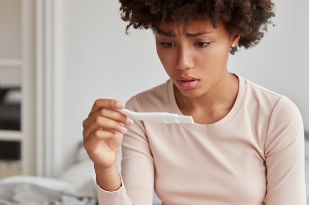 Verlegen vrouw met Afro-kapsel staat voor een ernstig probleem, ontevreden over de zwangerschapstest