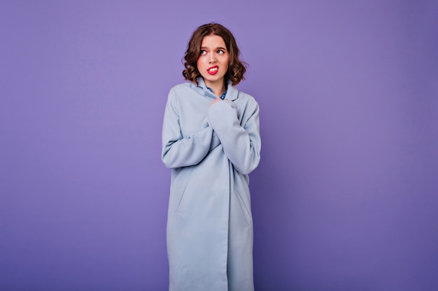 Verlegen vrouw met kort golvend haar poseren in elegante lange jas. mooi europees krullend meisje in blauwe kledij geïsoleerd op paarse muur.