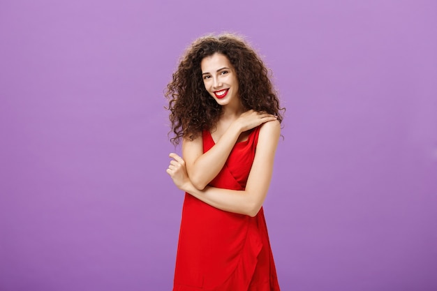 Verlegen vrouwelijke elegante europese vrouw met krullend kapsel en lippenstift in rode luxe jurk touchi...
