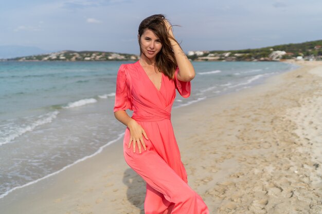 Verleidelijke brunette vrouw met tatoo bij de hand in roze jurk poseren op het strand.