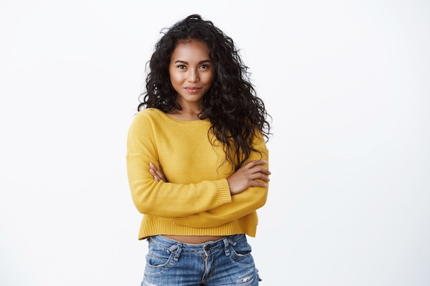 Verleidelijke jonge Afro-Amerikaanse vrouw met donker krullend kapsel, vastberaden en gemotiveerd glimlachen, zelfverzekerde pose met kruisarmen op de borst, lachende gedurfde camera, witte muur
