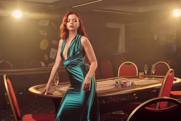 Verleidelijke roodharige meid in een lange blauwe satijnen jurk poseert zijwaarts tegen een pokertafel en kijkt naar beneden in een luxe casino. passie, kaarten, chips, alcohol, winnen, gokken - het is als vrouwelijke enterta