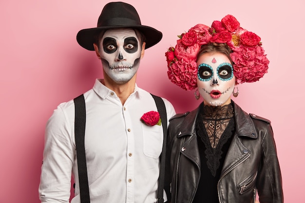 Verliefde paar in kostuums van skeletten en schedelmake-up, hebben uitdrukkingen bang gemaakt, herfstvakantie vieren, poseren tijdens horrorfeest, geïsoleerd op roze achtergrond. Gelukkig Halloween-tijdconcept