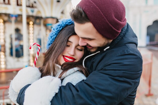 Verliefde paar omarmen in pretpark in winterweekend. Gelukkig zwartharige meisje kerst vieren met vriendje en poseren voor carrousel in koude dag.