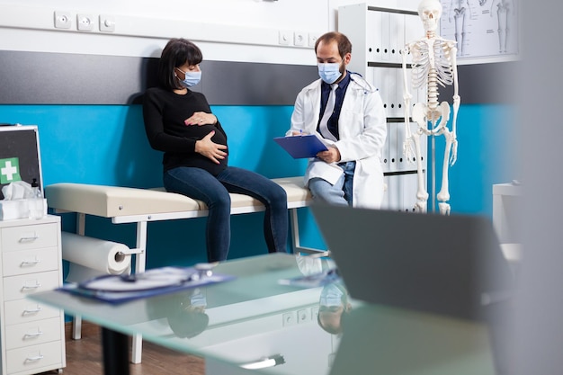 Verloskundige die aanstaande patiënt raadpleegt bij controleafspraak. Huisarts en zwangere vrouw doen medisch onderzoek, bespreken bevallingsondersteuning in kabinet.