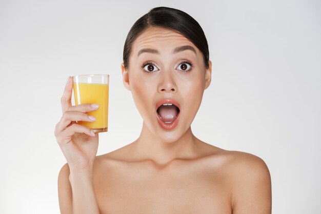 Vermakelijke foto van grappige vrouw met donker haar in broodje dat transparant glas vers-gedrukt jus d'orange houdt, dat over witte muur wordt geïsoleerd