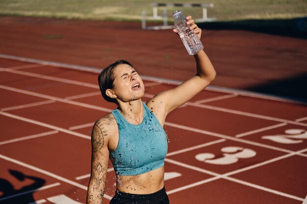 Vermoeid atletenmeisje in sportkleding die zoet water over zichzelf giet na het rennen op het stadsstadion