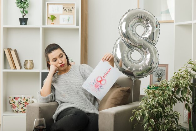 Vermoeid mooi meisje op een gelukkige vrouwendag die een wenskaart vasthoudt en bekijkt terwijl ze op een fauteuil in de woonkamer zit