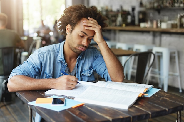 Vermoeide Afro-Amerikaanse student met krullend kapsel dat zijn wangen uitblaast en er verveeld of beu uitziet, geduld verliest terwijl hij geen ingewikkeld wiskundig probleem oplost, thuisopdracht doet